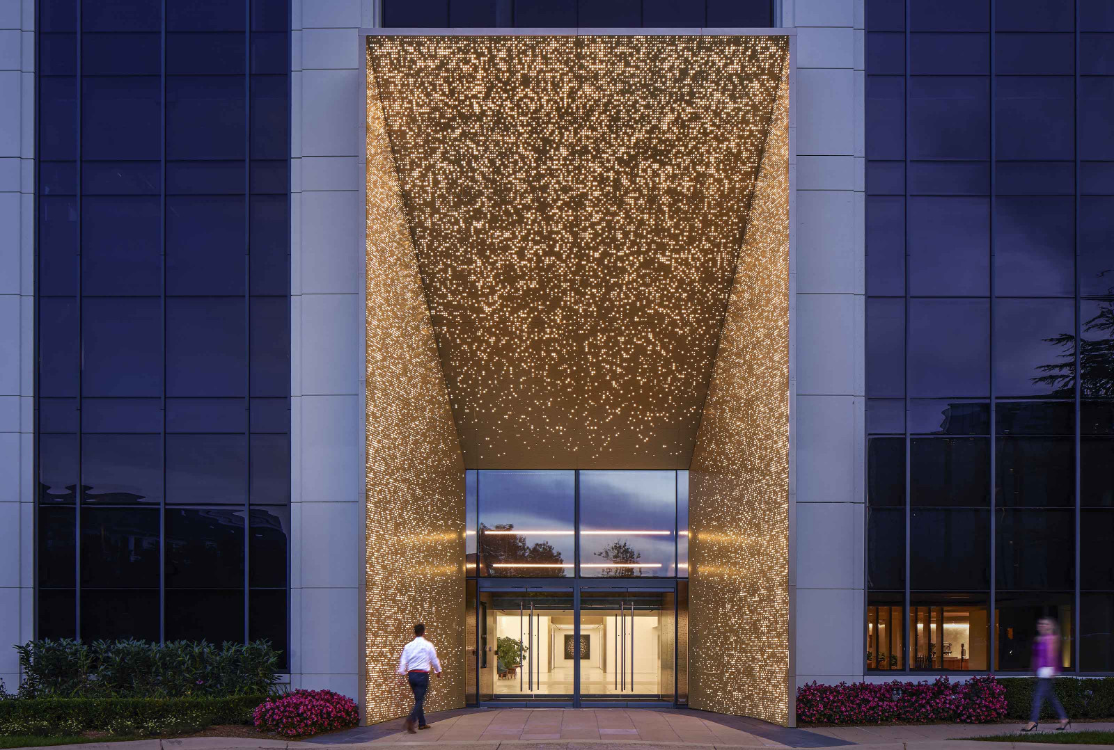 Exterior view of a commercial building with gold backlighting at its entrance. There is a person wearing a white shirt and black pants entering the building.