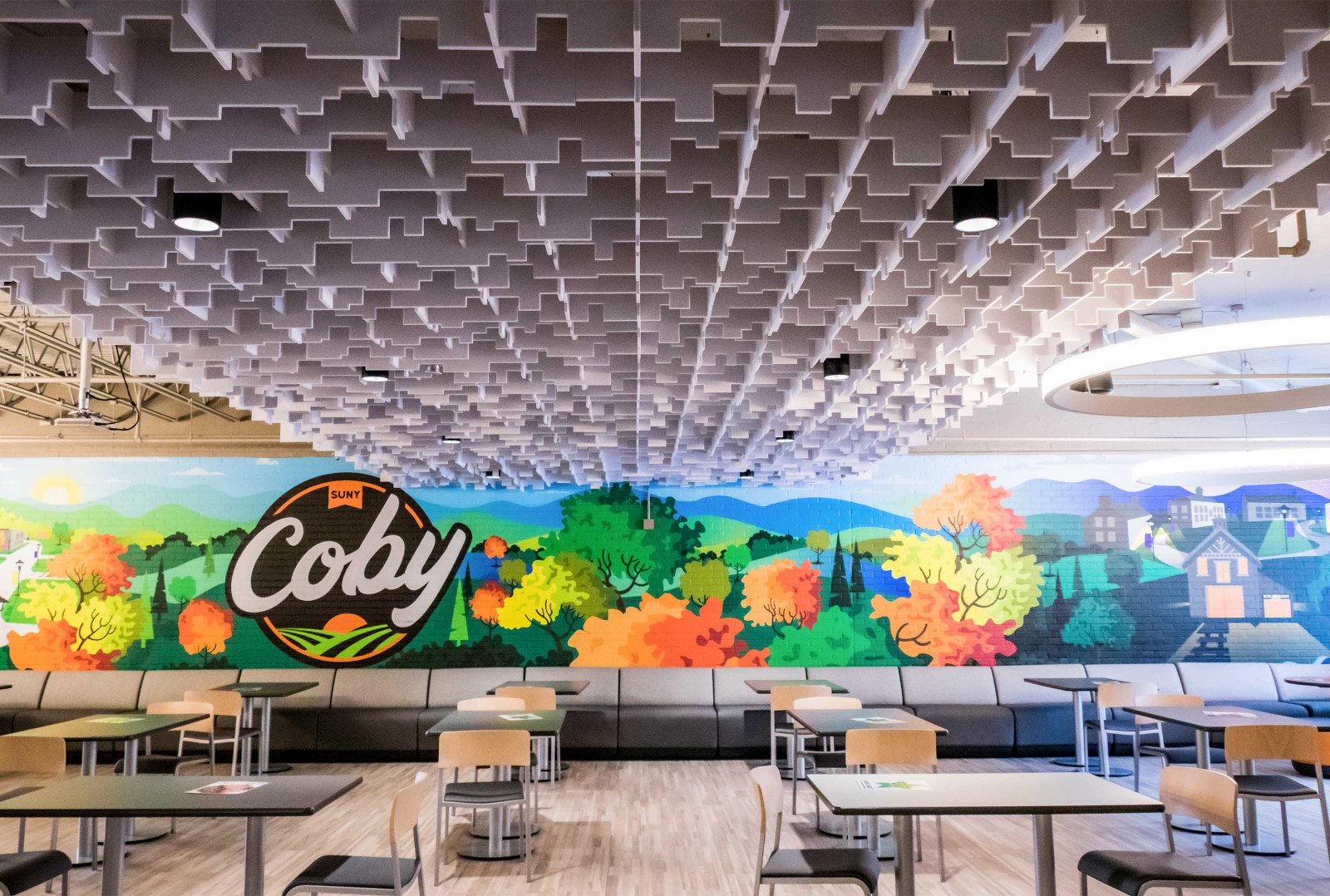 White SkyLine paneling lines the ceiling. There is a colorful mural with Coby’s company logo, trees and houses. A combination of booths and tables with chairs line the floor.