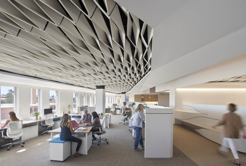 A white ceiling with weave-like paneling lines an office. The room is full of people at desks working. The bottom right corner contains a person mid-walk.