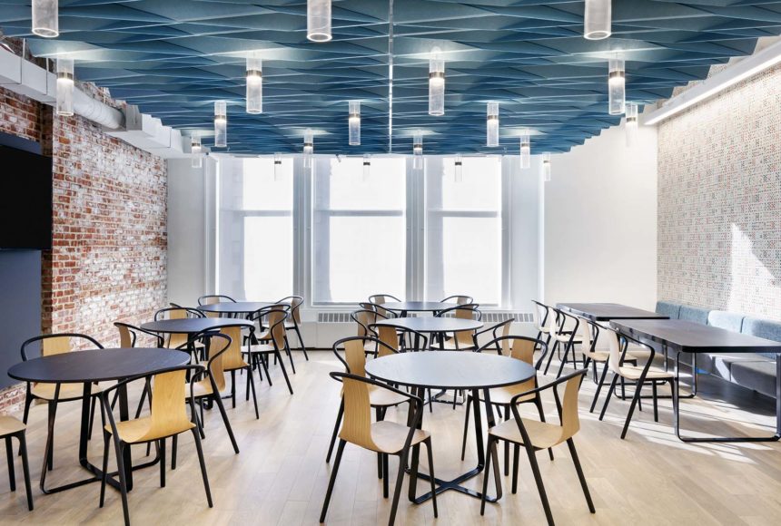 This room has blue weave-like paneling mixed with light fixtures. The room is full of tables and chairs. There are three windows and a brick wall.