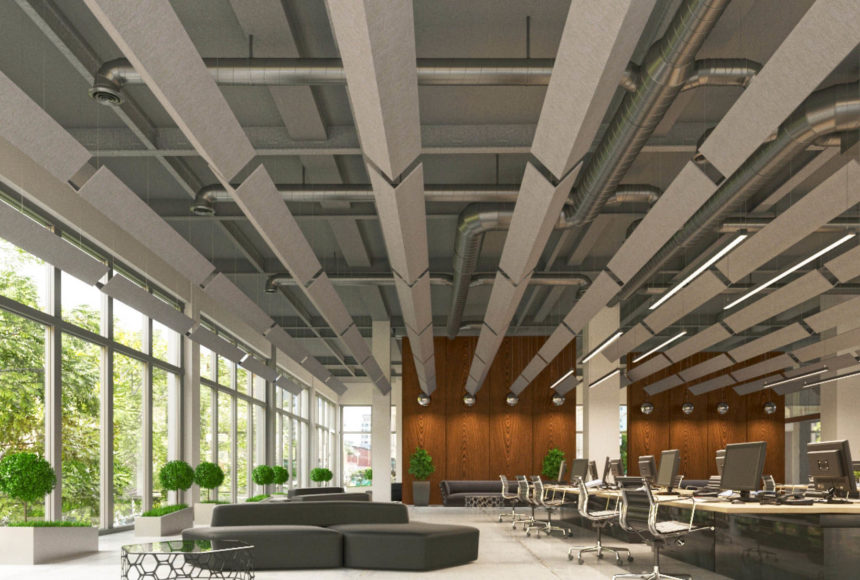 Metal ductwork and pointed rows of grey paneling make up the ceiling. Windows and rounded plants line the left wall while a long table with monitors and desk chairs lines the right side. 