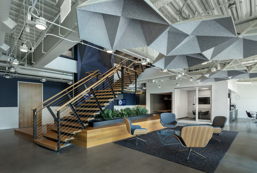 There are steps in the center of the room with a set of four chairs and a rectangular planter. White ductwork and grey hexagonal ceiling panels make up the ceiling. 