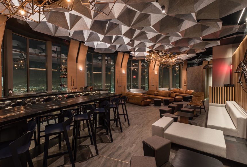 Gathering area with dark lighting and acoustic baffles on the ceiling. Various seating options are available, and a view of the city is seen through large windows.