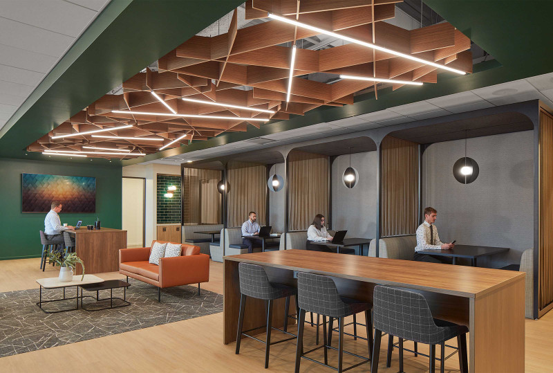 This office space features wood tables, black chairs, a leather sofa and four work booths. There are Arktura baffles above the space with bright lighting.  