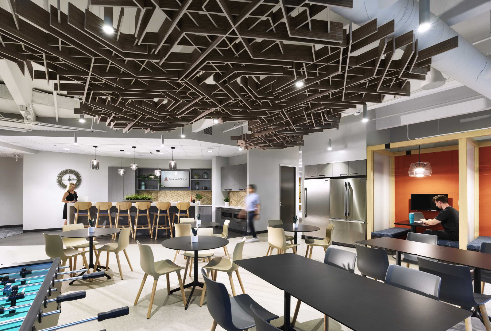 A breakroom at the Carlyle Amenity Center, featuring several booths on the right-hand side, several black tables in the center, a foosball table on the left and a small kitchen in the back. SoftGrid Flux panels with a wood texture are hanging from the ceiling.