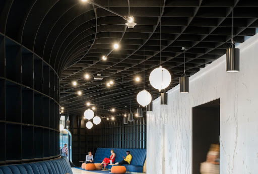 Black curved panels start on the left wall and continue onto the ceiling. There are ball-like light fixtures combined with hanging can lights. People are sitting on a blue couch with tables and ottomans.